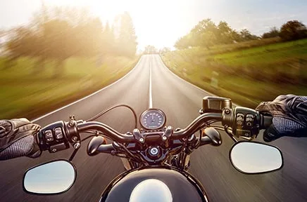 Blick vom Motorrad auf eine sonnige Landstraße mit Lenker, Tachometer und Spiegeln im Vordergrund, umgeben von grünen Feldern und Bäumen.