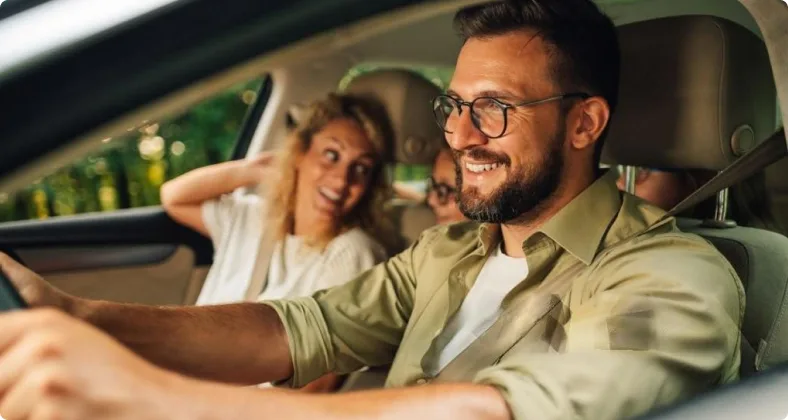 Ein lächelnder Mann mit Brille fährt Auto, während im Hintergrund eine fröhliche Frau als Beifahrerin zu sehen ist.